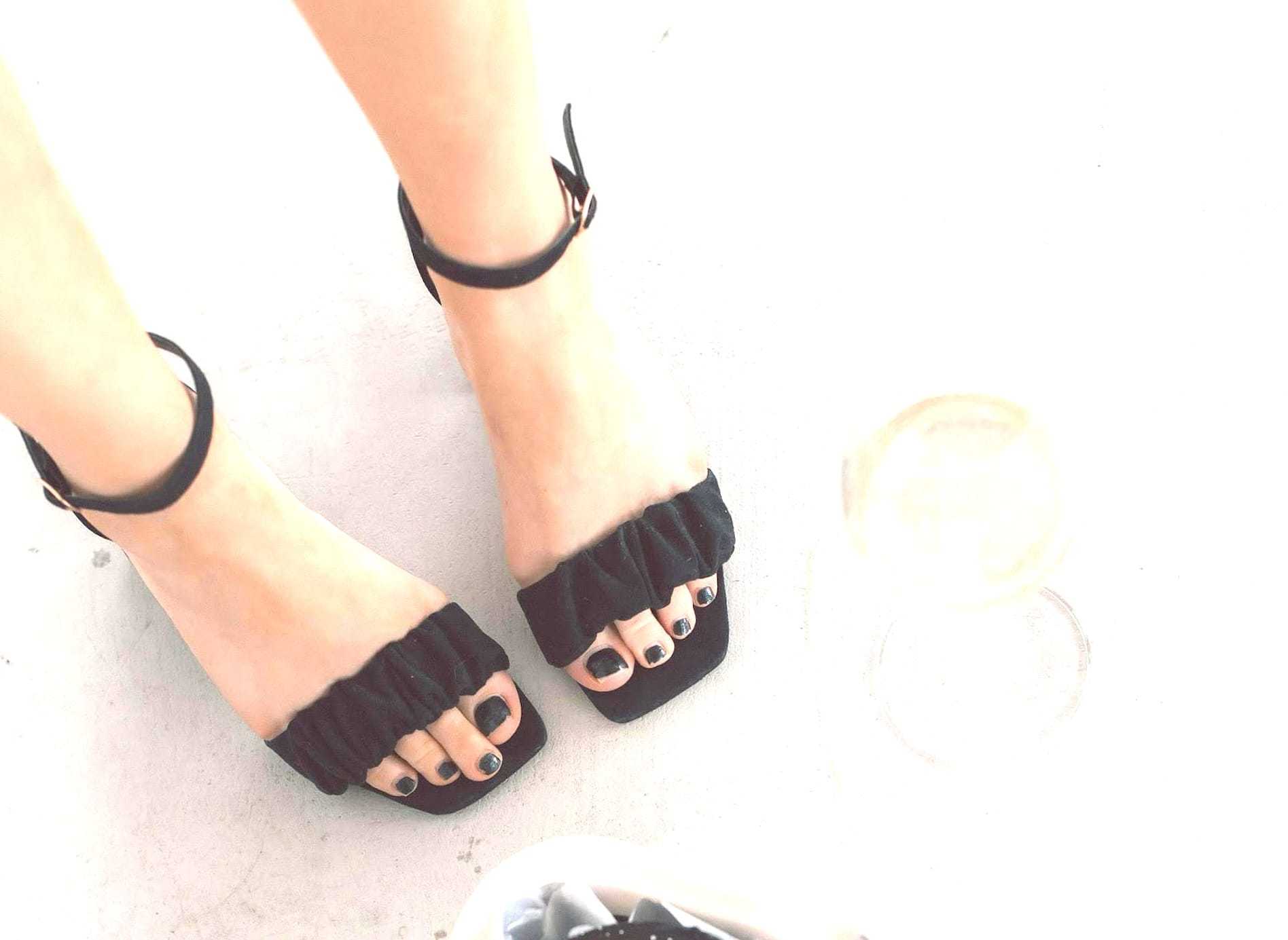 Woman wearing black open-toe heels with dark nail polish, standing on a light-colored floor.